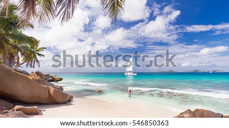 Woman wearing long floral summer dress and hat waving to people on the catamaran on Anse Patates beach, La Digue Island, Seychelles. Summer vacations on picture perfect tropical island concept.