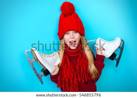 Cheerful little girl in warm sweater and hat holding figure skates. Blue background. 