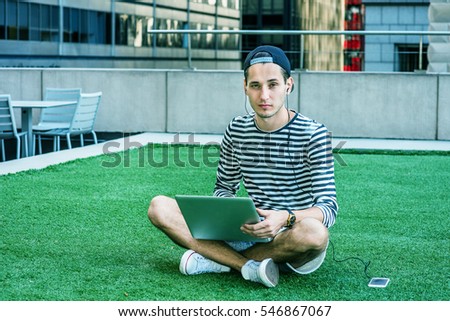 Russian man studies in New York, wearing striped long sleeve T shirt, cap worn backward, crossing legs, sits on green lawn, works on laptop computer, listens music on cell phone. Color filtered effect