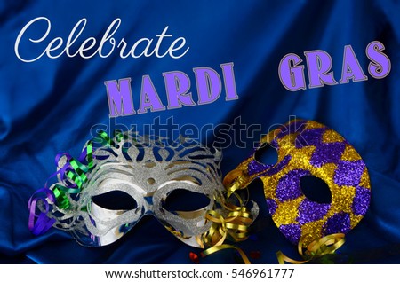 Silver feminine Mardi Gras mask with masculine harlequin mask with curled ribbons and colorful confetti on a luxurious blue satin draped background. Mardi Gras, meaning Fat Tuesday, text added