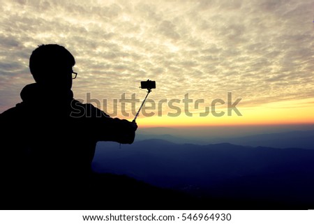 silhouette of a man taking a picture with a selfie stick on top of mountain before a sky sunset background
