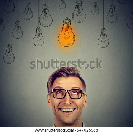 Portrait happy handsome man in glasses looking up at bright idea light bulb above head isolated on gray wall background