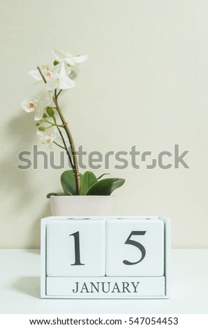 Closeup white wooden calendar with black 15 january word with white orchid flower on white wood desk and cream color wallpaper in room textured background , selective focus at the calendar