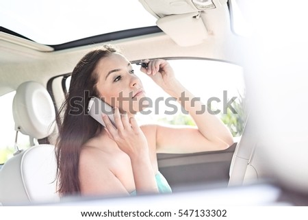 Young woman using mobile phone while applying mascara in car