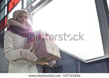 Smiling woman in warm clothing carrying stacked gifts by window