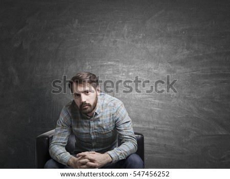 Portrait of a young man with a beard. He is sitting in a leather armchair and wearing a checkered shirt. A blackboard is in the background. Mock up