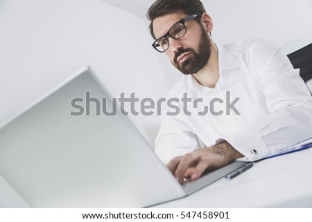 Tilted portrait of a young bearded boss working in his office. He is wearing glasses and looking at his laptop screen while typing