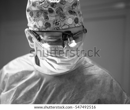 Dramatic black and white photo of the operating surgeon in the surgery room. Surgeon in mask and glasses with mounted headlight. Close portrait
