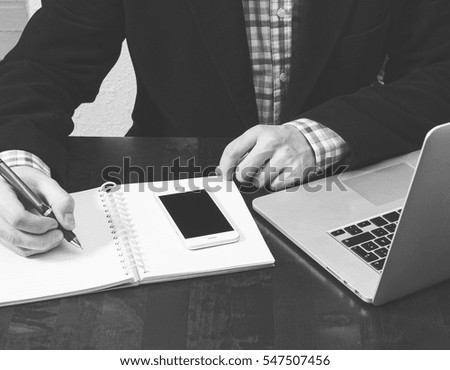 Business concept image. Business man is writing a memo. Mobile phone and a laptop on the wooden table. Business outfit. Image in black and white.