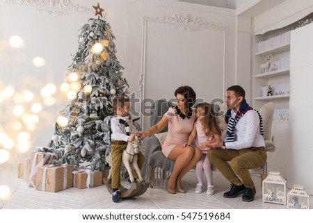 New Year. Beautiful and happy family, mother, father, son and daughter. Parents sit on the sofa near the Christmas tree, a boy riding a wooden horse. Under the Christmas tree gifts, shine lights.
