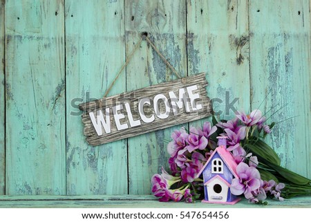 Wood welcome sign by birdhouse and purple spring flowers hanging on antique rustic mint green wooden background; springtime background with copy space