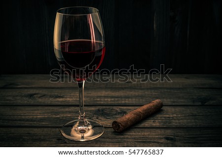 Glass of red wine and cuban cigar on an old wooden table. Focus on the cuban cigar, image vignetting and the orange-blue toning