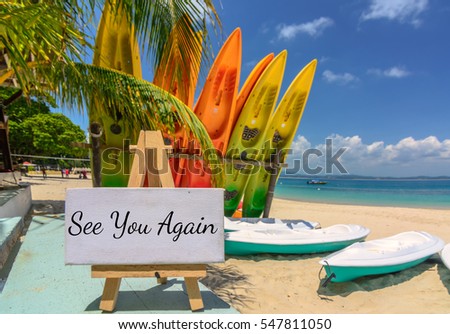 Conceptual image with word "SEE YOU AGAIN" on white canvas frame and wooden tripod. Blurred image of beach and colorful kayaks background. Image with selective focus
