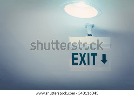 White sign with word exit and arrow pointing down hanging on the white ceiling with lamp. Toned