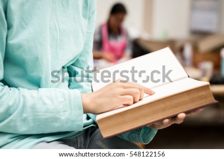Asian young girl close up finger pointing to reading a book at outdoor at home, relax time with her parent blurry background.