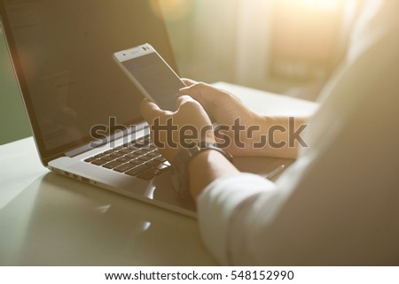 Silhouette of cropped shot of a young man working from home using smart phone and notebook computer, man's hands using smart phone in interior, man at his workplace using technology, flare light.