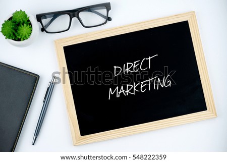 Top view of diary, pen, eye glasses, plant and black chalkboard written with DIRECT MARKETING on white background.
