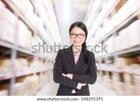 Young Asian female store manager arms crossed standing in warehouse. Shelves with goods at background.