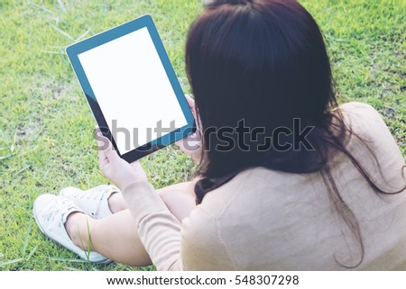 Mockup image of an asian woman sitting and holding black tablet with blank white screen on green grass at the park