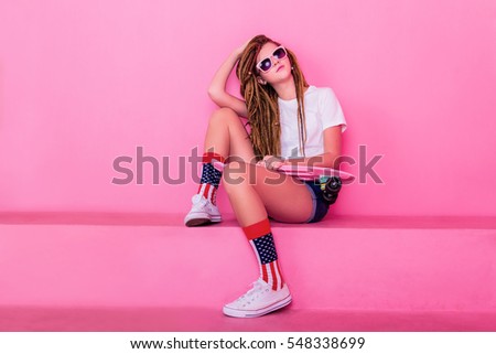 Pretty girl in sunglasses with dreadlocks sitting holding pink skateboard skateboard