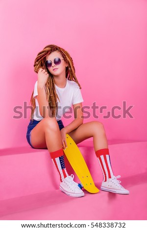 Pretty girl in sunglasses with dreadlocks sitting near the yellow skateboard