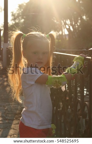 Little girl on roller skates with protective pads poses near railing in sunny park