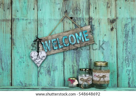 Welcome sign with silver tin heart hanging over glass jelly jars on antique rustic mint green wood background; home and love concept background with painted copy space