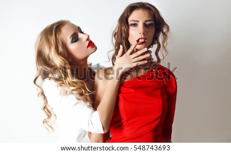 Two young girl friends fighting,having a quarrel.combat,flirt,relationships,lips,touching lips,blonde and brunette.One keeping brunette's hair. Second pulling blonder's wear. Casual style.white wall
