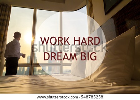 Maid-up bed in cozy room. Young businessman with coffee cup standing at window looking at city scenery on the background. Motivational text "Work hard Dream big"