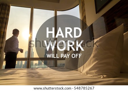 Maid-up bed in cozy room. Young businessman with coffee cup standing at window looking at city scenery on the background. Motivational text "Hard work will pay off"