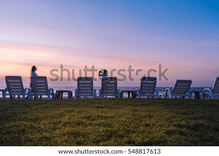 Woman takes pictures on the phone at sunset near the sea and and beach chairs seen in the foreground
