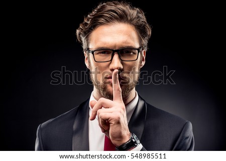 Handsome businessman in eyeglasses and suit gesturing for silence with finger isolated on black Royalty-Free Stock Photo #548985511