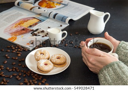Cup of coffee in woman hands. Saucer with cookies, roasted coffee beans and culinary magazine on dark table