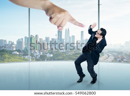 A scared businessman  in a scare pose and a giant hand pointing at him, on the office background. Business and management, employment issues or getting fired.  Royalty-Free Stock Photo #549062800