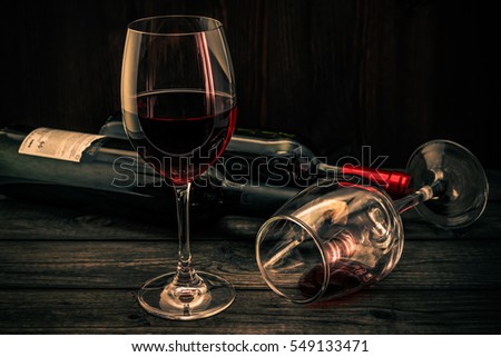 Two bottles of red wine with two glasses on an old wooden table. Focus on an overturned glass of wine, image vignetting and the orange-blue toning