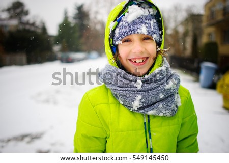 Portrait of the schoolboy walking in winter day. The boy is dressed in a bright jacket and a knitted cap and a scarf. His face and clothes are closed up with snow. The child happily smiles.