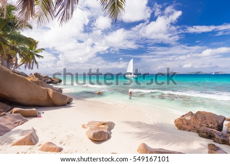 Woman wearing long floral summer dress and hat waving to people on the catamaran on Anse Patates beach, La Digue Island, Seychelles. Summer vacations on picture perfect tropical island concept.
