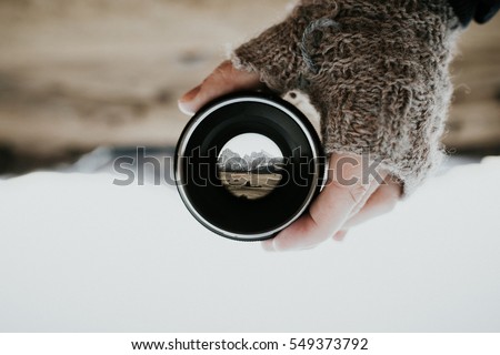 A mountain range photographed through a camera lens held by a human hand with gloves. The picture is turned upside down. A shallow depth of field creates a very prominent background blur.