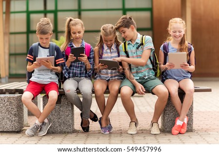 primary education, friendship, childhood, technology and people concept - group of happy elementary school students with backpacks sitting on bench and talking outdoors Royalty-Free Stock Photo #549457996