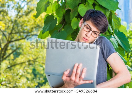 Asian American college student studies in New York. Man wearing glasses, stands by green big leaf plants, works on laptop computer, talks on phone held between head and shoulder.