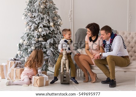 Christmas. Beautiful and happy family, mother, father, son and daughter. Parents sit on the sofa near the Christmas tree, a boy riding a wooden horse. Under the Christmas tree gifts, shine lights.
