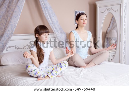 Happy mother and daughter meditating and smiling while sitting in yoga pose on the bed at home. Studio shot.