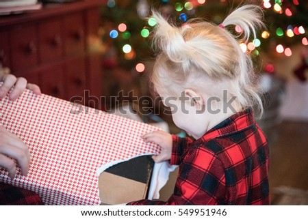 Little 2 year old blonde girl with pigtails and red plaid buffalo check pajamas unwrapping a present on Christmas Day morning with blurry multicolored lights bokeh in background - soft focus