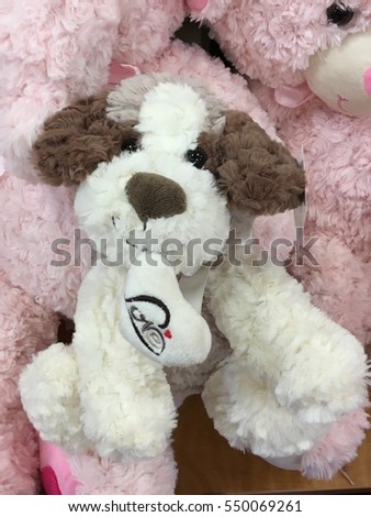 A picture of stuffed  toy dog is sitting in the middle of pink background showing white body, big  black eyes, brown ears and nose, looking straight at you with heart shaped love letter in his mouth
