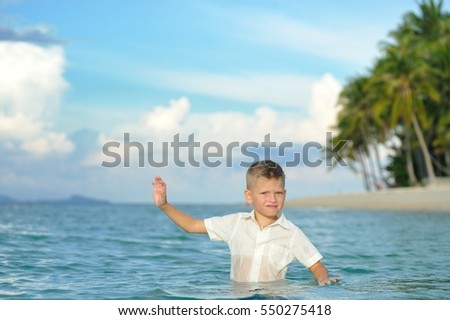 Waist portrait on a tropical beach: slim and beautiful 8 years old boy in clothes standing in water. His pants are already wet, and shirt gets wet from the splashing