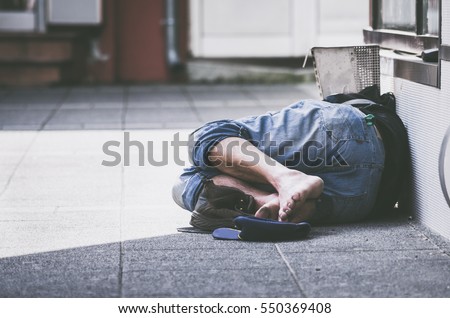 Homeless man sleeps on the street in the shadow of the building.  Royalty-Free Stock Photo #550369408