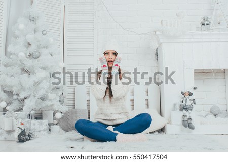 the girl dressed in warm sweater, hat, and gloves sits near the fireplace and Christmas tree .