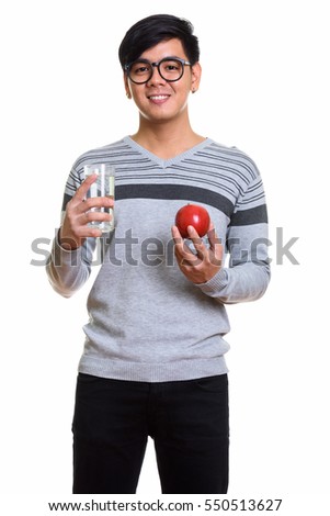 Studio shot of happy Asian man smiling while holding red apple and glass of water isolated against white background