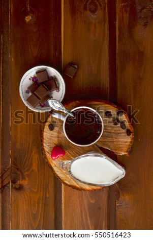 a cup of coffee next to the chocolate pieces standing on a tree saw cut on a brown wooden table near full creamer