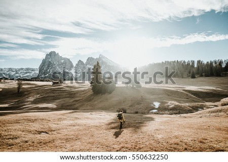 A person in a yellow Jacket walking over a field in the mountains in northern italy. The sunrise is creating heavy back light.